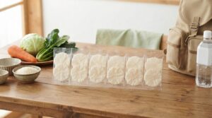 赤ちゃんから高齢者までを想定した長期保存用の米せんべいをテーマにした食品ブログのアイキャッチ画像。明るい木製テーブルの上に、口どけのよさそうな白い薄焼き米せんべいを2枚ずつ透明な個包装にし、6袋並べる。横に国産米、にんじん、キャベツ、ほうれん草、防災リュック、保存水を控えめに配置する。白、淡い緑、ベージュを基調にした清潔感のある写実的な写真。横長16:9。実在の商品パッケージ、企業ロゴ、ブランド名、文字、乳幼児、高齢者などの人物は入れない。長期保存できるやさしい米菓だと伝わる構図。